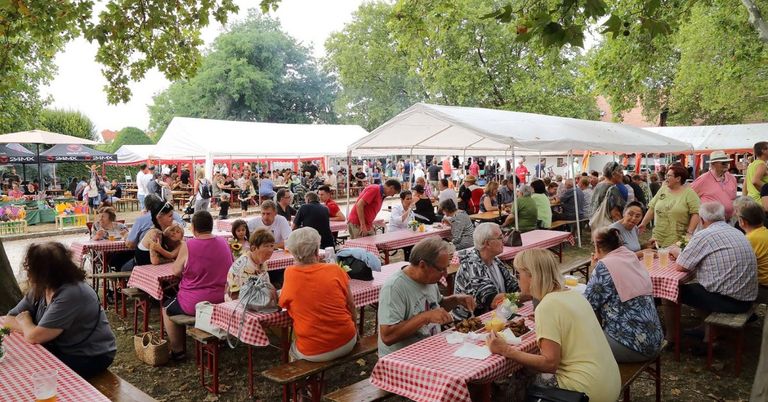 Eine große Gruppe von Menschen sitzt an Tischen unter weißen Zelten in einem Außenbereich und genießt Essen und Getränke. Bäume und ein Gebäude sind im Hintergrund zu sehen.