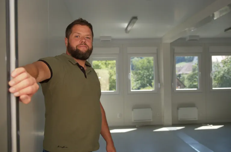 A man with a beard wearing a green polo shirt is standing in an empty room with white walls and windows.