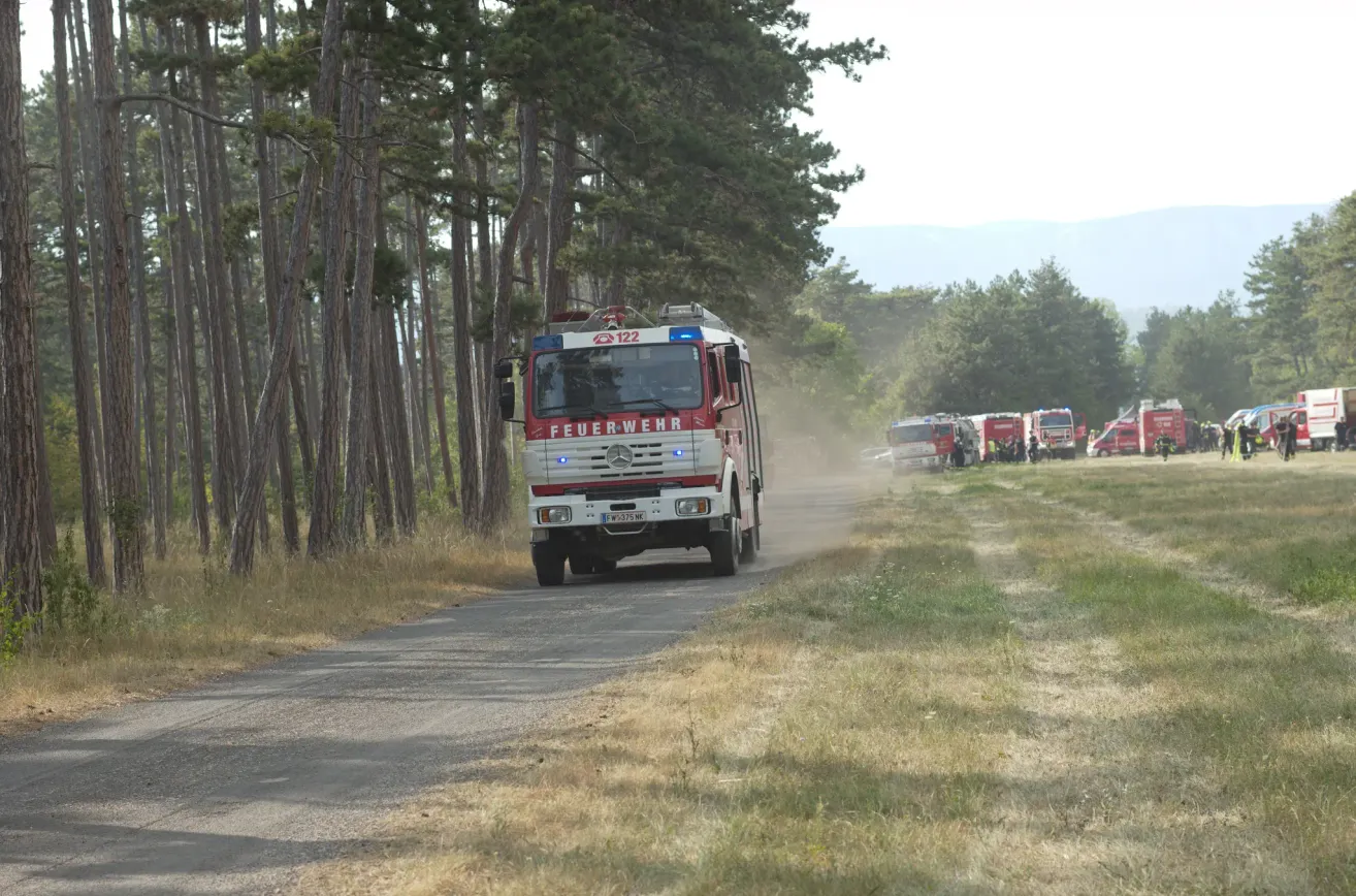 Ein roter Feuerwehrwagen mit der Nummer 122 fährt auf einer Schotterstraße umgeben von Bäumen. In der Ferne sind andere Feuerwehrwagen geparkt.