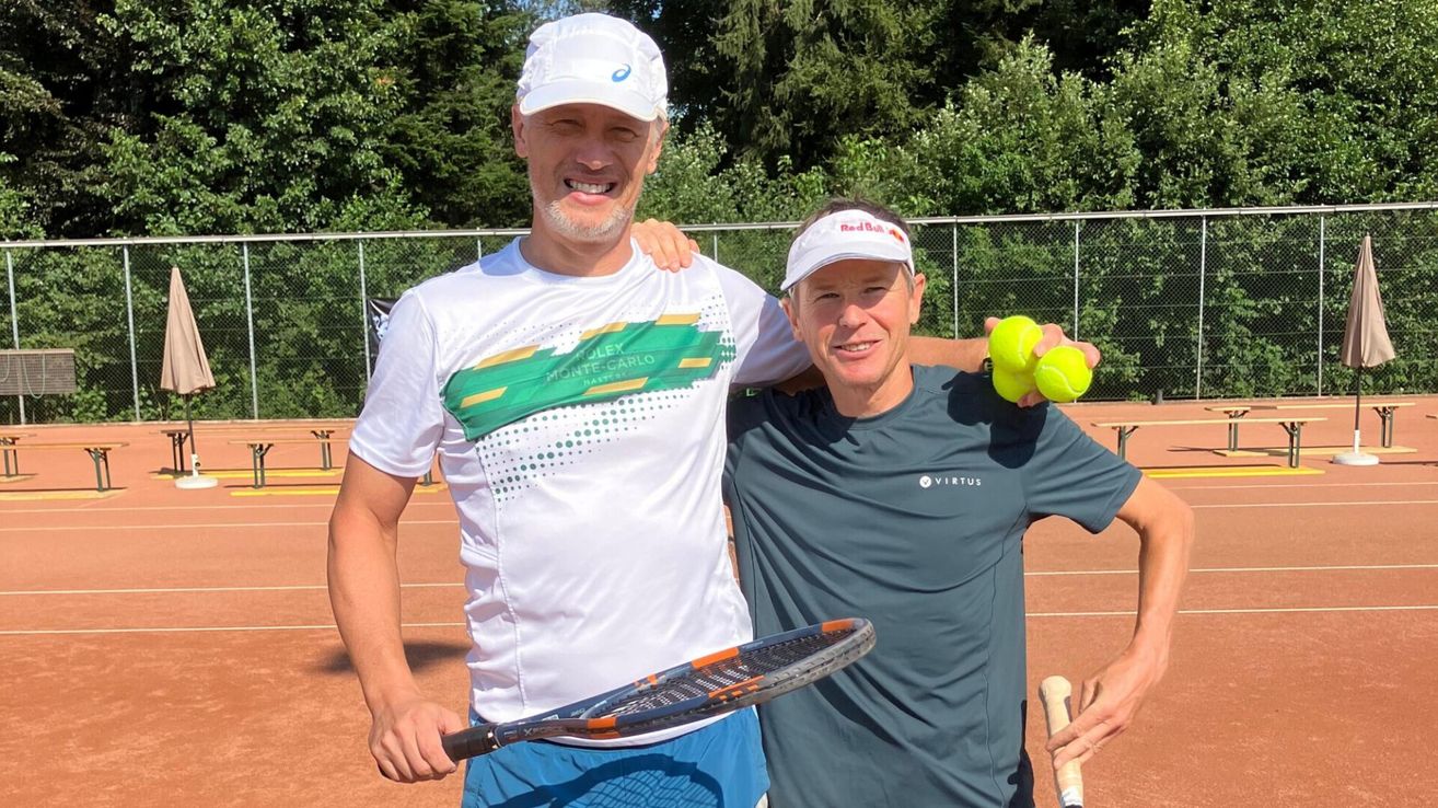 Zwei Männer stehen auf einem Tennisplatz, einer hält eine Tennisschläger und der andere hält Tennisbälle. Sie lächeln beide und scheinen für ein Foto zu posieren.