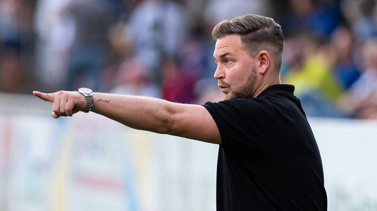 Ein Mann mit einer Tätowierung am Arm, der ein schwarzes T-Shirt trägt, gestikuliert mit der rechten Hand. Er scheint ein Trainer oder Athlet zu sein.