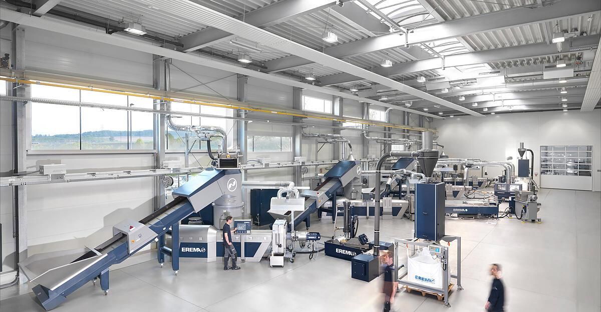 Eine Industrieanlage mit großen Maschinen und Fördersystemen. Arbeiter bedienen und überwachen die Geräte. Der Raum ist gut beleuchtet mit Deckenleuchten und großen Fenstern.