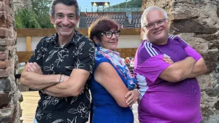 Drei Menschen stehen vor einer Ziegelmauer und lächeln für ein Foto. Der Mann links trägt ein schwarzes Blumenhemd, die Frau in der Mitte eine blaue Robe und der Mann rechts ein lila Shirt. Hinter ihnen ist ein Holzzaun und ein Stadion zu sehen.