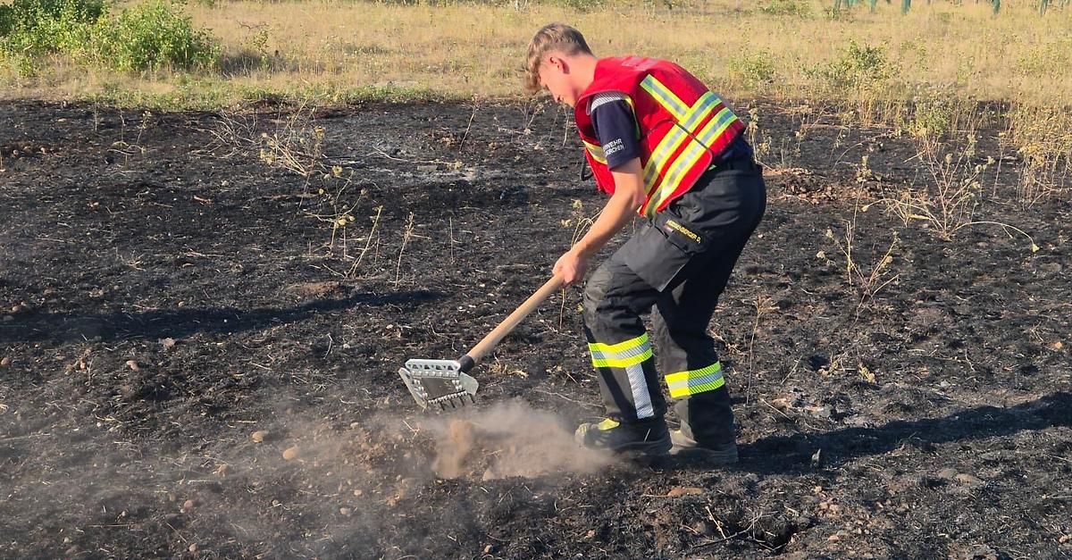 Ein Feuerwehrmann in einer reflektierenden Weste und Hose mit reflektierenden Streifen räumt mit einem Rechen Asche von einem verbrannten Feld.
