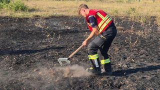Ein Feuerwehrmann in einer reflektierenden Weste und Hose mit reflektierenden Streifen räumt mit einem Rechen Asche von einem verbrannten Feld.