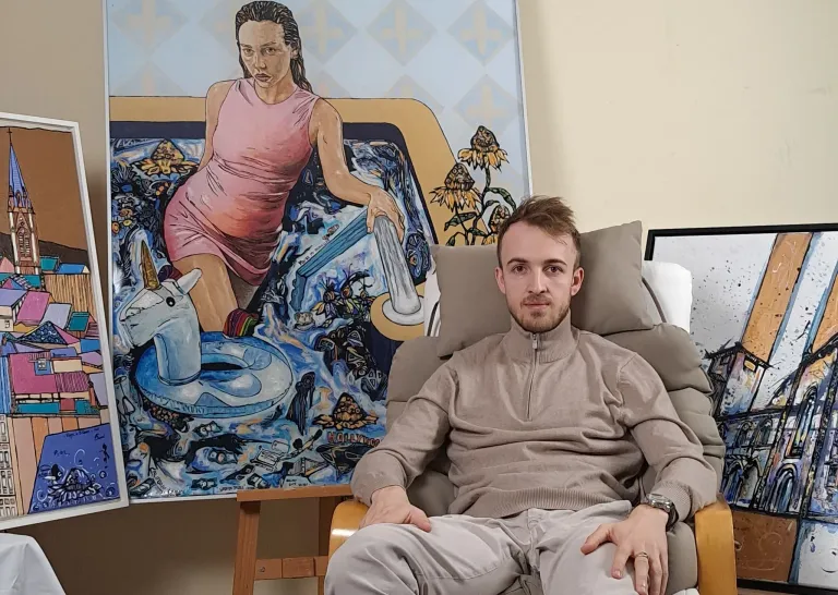 A man sits on a chair in front of a large painting of a woman in a pink dress in a bathtub with a unicorn and floating objects.