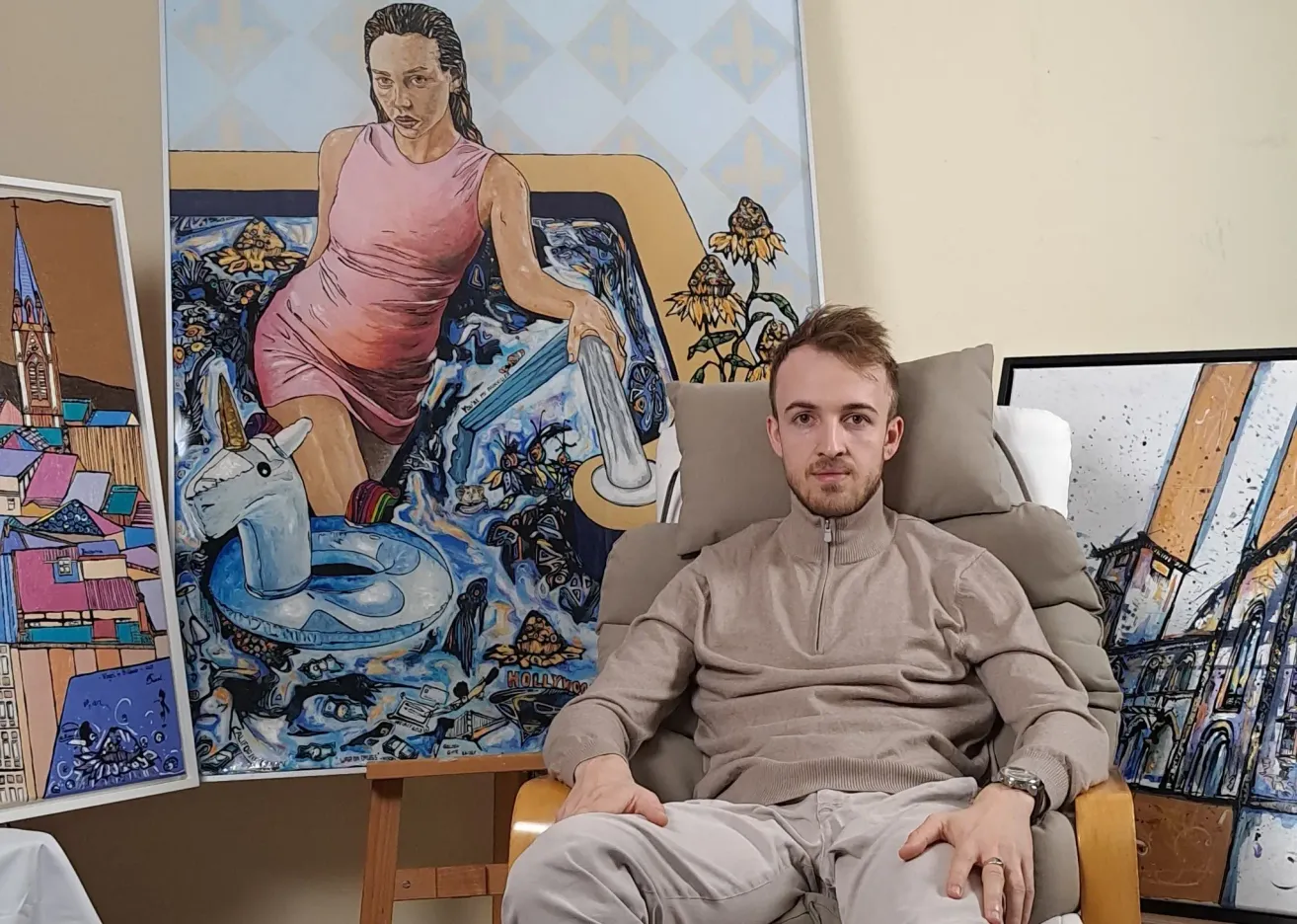 A man sits on a chair in front of a large painting of a woman in a pink dress in a bathtub with a unicorn and floating objects.