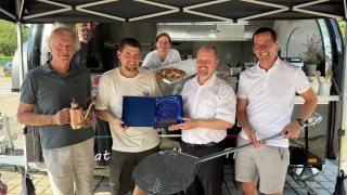 Four men are standing under a tent, holding a pizza peel and a blue award. They are smiling and posing for a photo. Behind them, a woman is holding a pizza box.