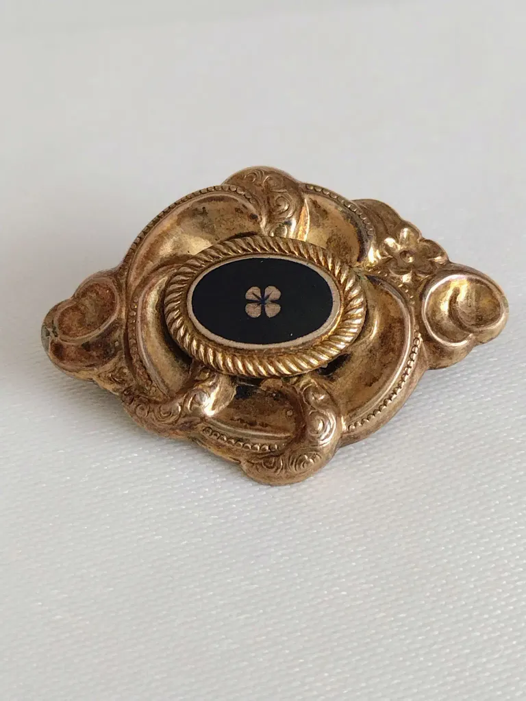 An antique gold brooch with an oval black center featuring a white four-leaf clover design, set against a textured gold background.