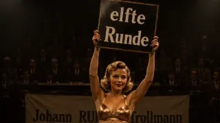 A woman wearing a gold bikini holds up a sign that reads 'elfte Runde' in front of a blurred audience.