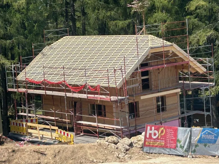 A wooden house under construction with scaffolding around it, located in a forested area. A red sign with white text is visible on the right side.
