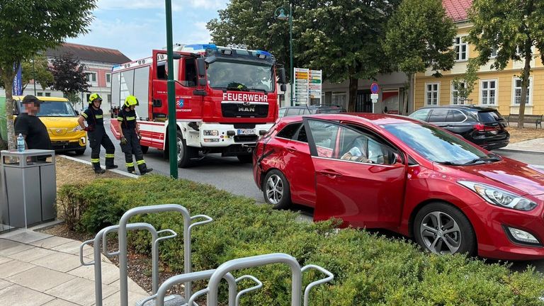A red car with its door open is parked on the side of the road. A firefighter is standing next to a red fire truck. Trees and a building are in the background.