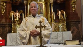 An elderly priest stands in front of an altar, holding a cross and wearing a white robe with golden embroidery. Behind him, two angels with candles are depicted on a golden background.