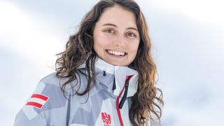 A smiling woman with curly brown hair wearing a gray and white jacket with a red logo on the left chest area.