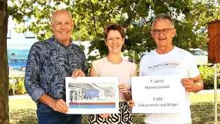 Three people stand under a tree, smiling and holding a poster that reads '9 Jahre Momentothek' and '9,000 Dokumente und Bilder.' The man on the left holds a framed picture, the woman in the middle holds a poster, and the man on the right wears a white shirt.