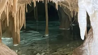 A cave with stalactites and stalagmites, water flowing through, and a narrow entrance.