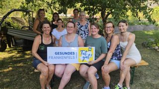 A group of women sit on a bench in a garden, holding a sign that reads 'GESUNDES FRANKENFELS Tut gut!' in German. They are all smiling and appear to be posing for a photo.