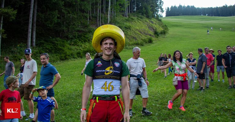 A man wearing a yellow bucket hat stands in a grassy field with a number 46 on his shirt, smiling. Behind him, several people are walking, including a woman with a handbag. Trees and a hill are in the background.