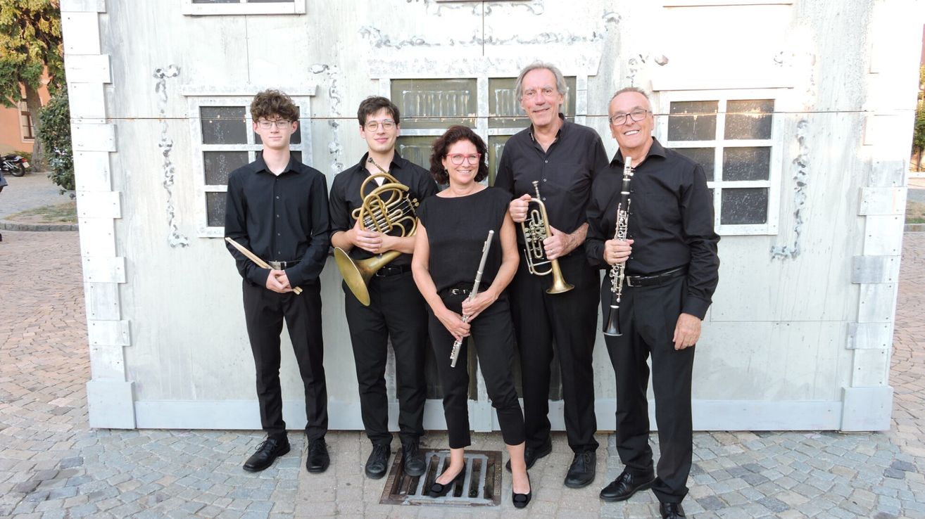 Fünf Musiker, alle in Schwarz gekleidet, stehen in einer Reihe und halten jeweils ein anderes Instrument in der Hand, lächeln für ein Foto. Hinter ihnen ist eine weiße Wand mit Glasfenstern.