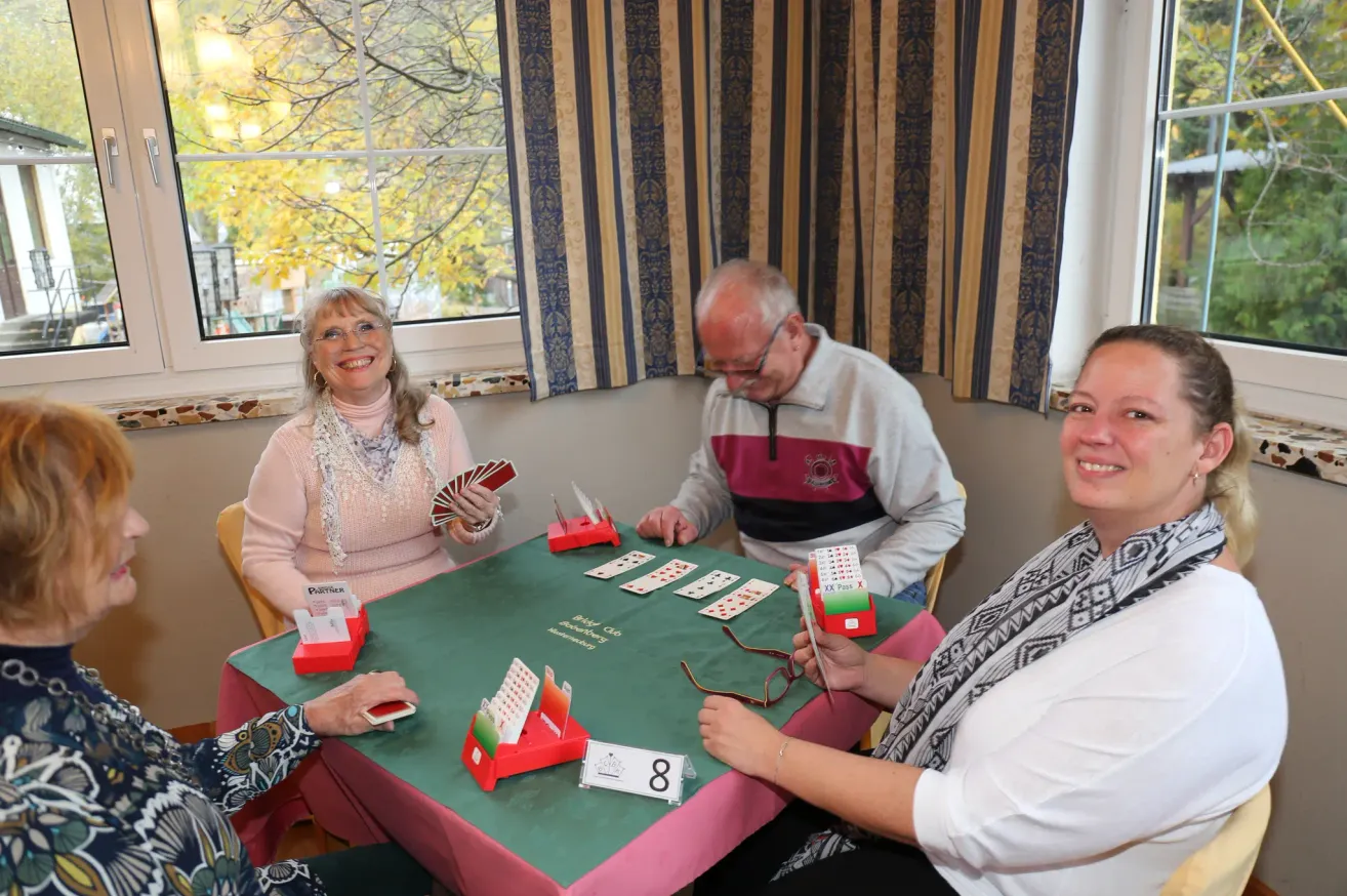 Four people are sitting around a table playing a card game. The table is covered with a green cloth and has cards and a red box on it. The woman on the left is smiling and holding a card. The man in the middle is looking at the cards on the table. The woman on the right is smiling and holding a card. Behind them is a window with curtains.