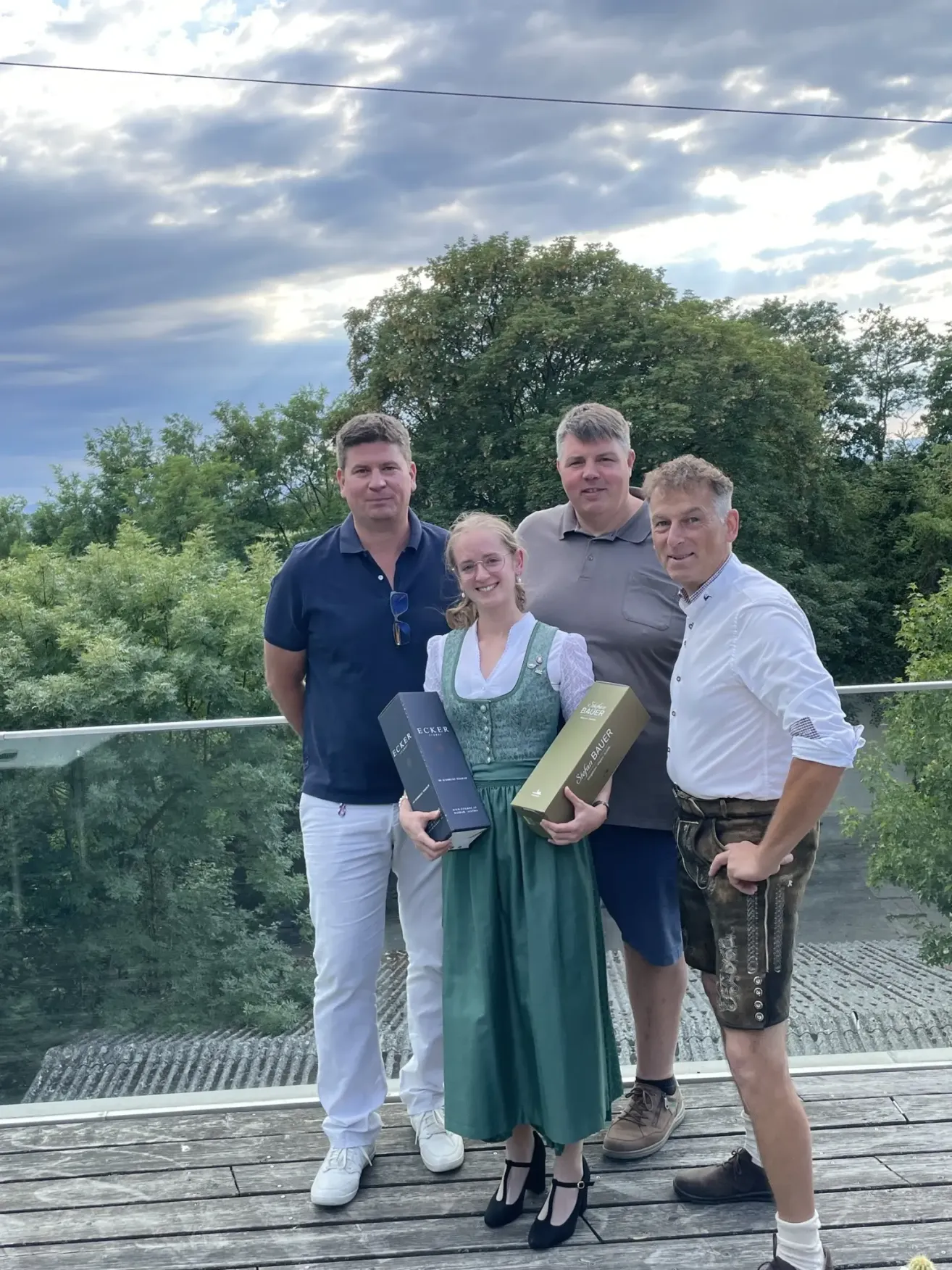 Four people are standing on a balcony, smiling for a photo. They are holding wine boxes. The woman is wearing a traditional dress. The background is filled with trees and a cloudy sky.