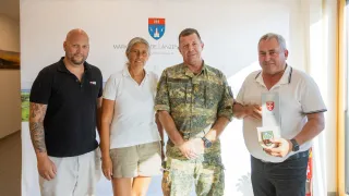 Four individuals are standing in front of a white backdrop with a logo. The man in the middle is in a military uniform, and the others are in casual attire. They are all smiling and appear to be posing for a photo.