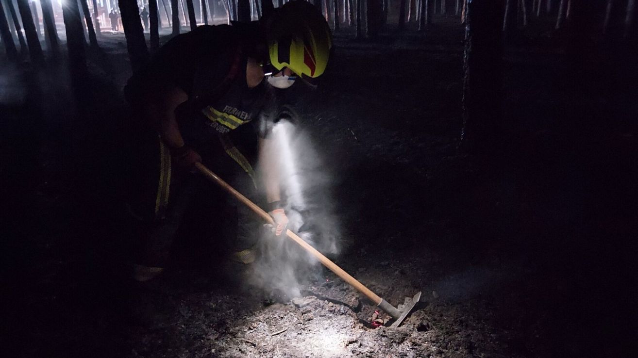 A firefighter in full gear, including a helmet and mask, uses a shovel to extinguish a fire in a forest at night.