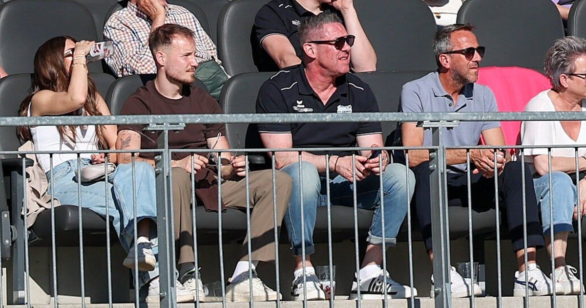 Four men sitting in a stadium, two wearing sunglasses, one in a black shirt with a logo, another in a brown shirt, and two others in blue and white shirts.