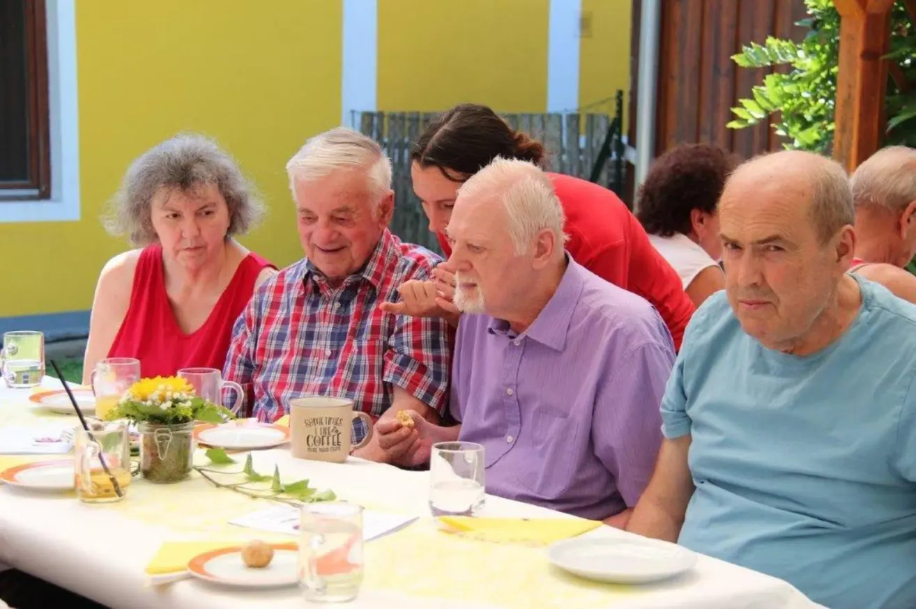 A group of elderly people sitting around a table, enjoying a meal. One man holds a coffee mug, and another has a sandwich. A woman in red stands behind them.