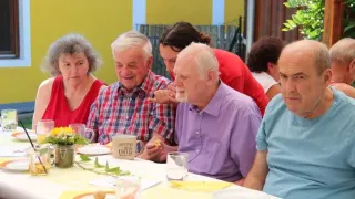 Eine Gruppe älterer Menschen sitzt um einen Tisch herum und genießt eine Mahlzeit. Ein Mann hält eine Kaffeetasse und ein anderer ein Sandwich. Eine Frau in Rot steht hinter ihnen.