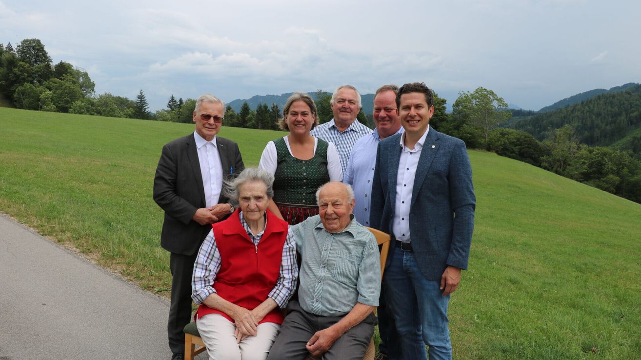 A group of people standing and sitting in a grassy field with mountains in the background.