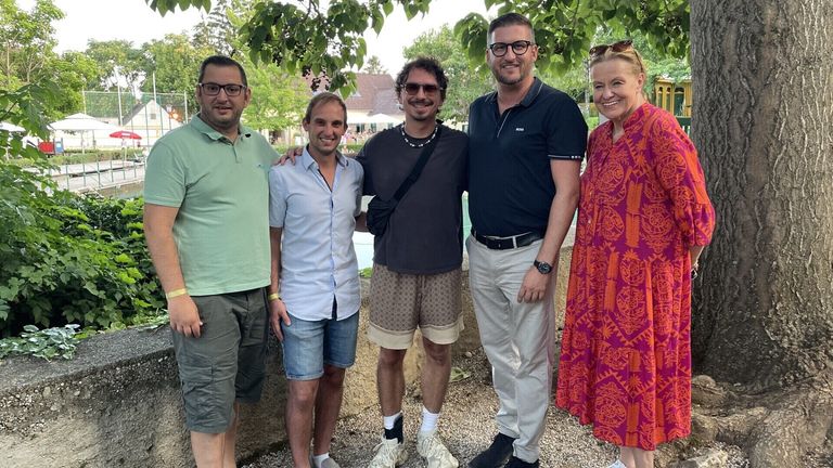 Fünf Personen stehen vor einem Gebäude mit einem Swimmingpool. Sie lächeln und posieren für ein Foto. Der Mann in der Mitte trägt eine Halskette und eine Sonnenbrille. Der Mann auf der rechten Seite trägt eine Uhr. Die Frau auf der rechten Seite trägt ein Armband.