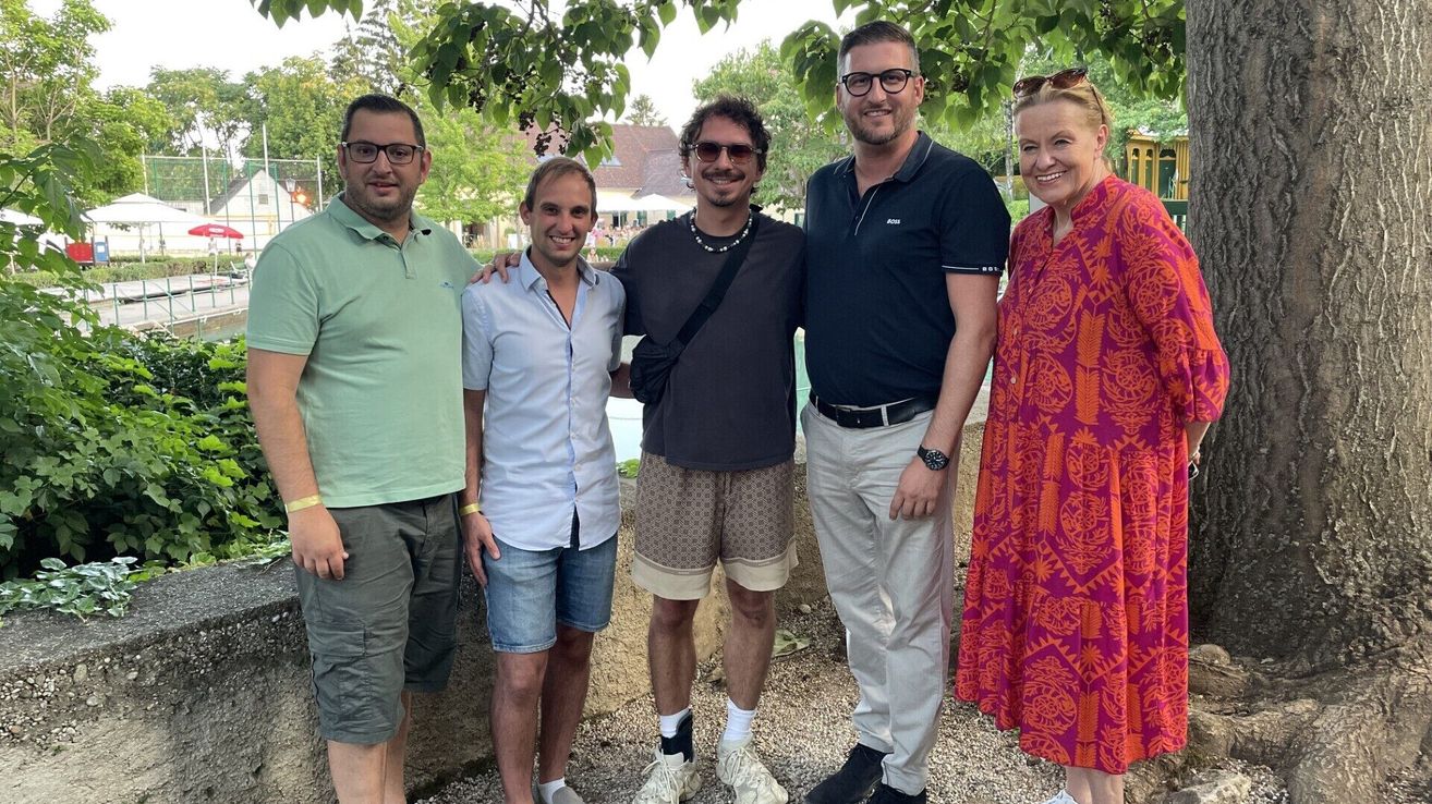 Fünf Personen stehen vor einem Gebäude mit einem Swimmingpool. Sie lächeln und posieren für ein Foto. Der Mann in der Mitte trägt eine Halskette und eine Sonnenbrille. Der Mann auf der rechten Seite trägt eine Uhr. Die Frau auf der rechten Seite trägt ein Armband.