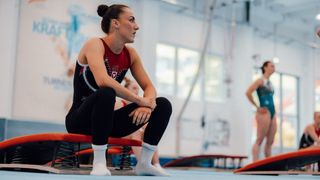 A female gymnast sits on a red spring board, wearing a red and black leotard, looking focused in a gym.