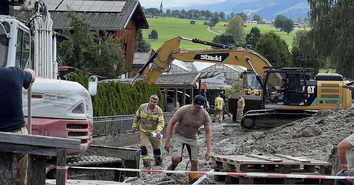 Two men in work uniforms are walking on a muddy road with a large excavator in the background.