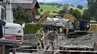 Two men in work uniforms are walking on a muddy road with a large excavator in the background.