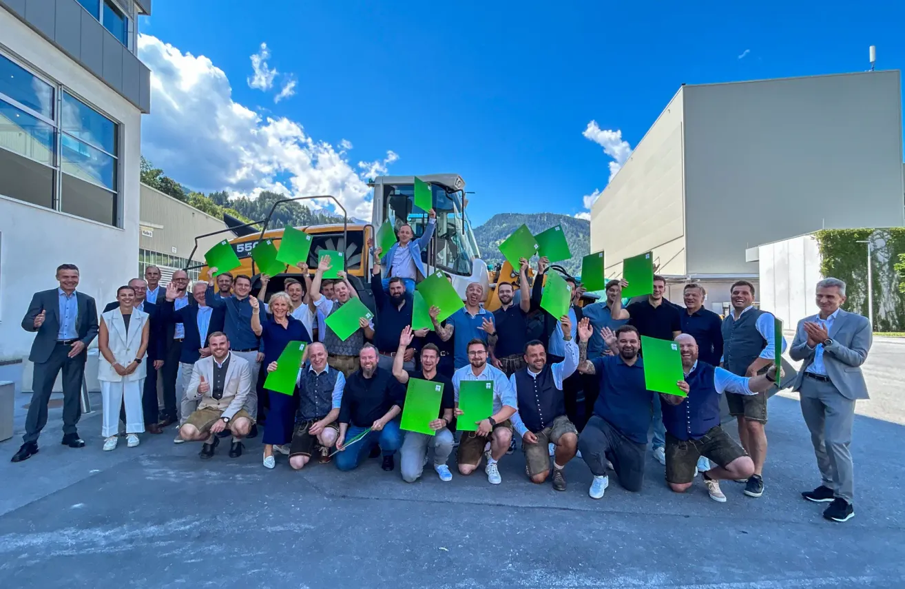 Eine Gruppe von Menschen posiert für ein Foto vor einem Baufahrzeug, hält grüne Papiere und lächelt. Im Hintergrund befinden sich ein Gebäude und Berge unter einem blauen Himmel mit Wolken.