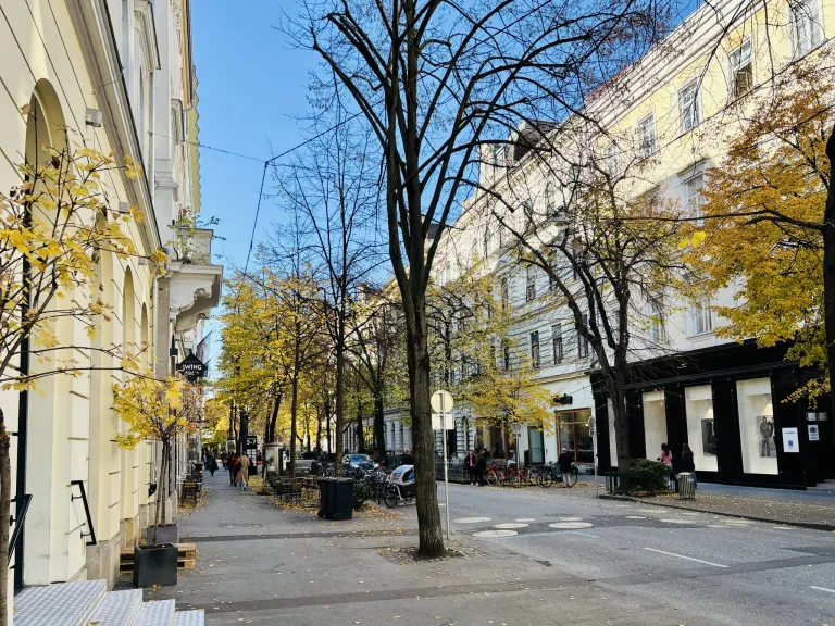Eine Straße mit Bäumen auf beiden Seiten. Menschen gehen auf dem Bürgersteig und einige fahren Fahrräder. Es gibt auch einige Mülleimer und ein Schild auf dem Bürgersteig.