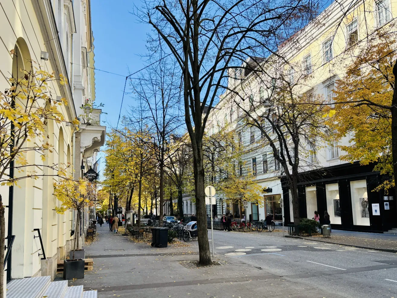 Eine Straße mit Bäumen auf beiden Seiten. Menschen gehen auf dem Bürgersteig und einige fahren Fahrräder. Es gibt auch einige Mülleimer und ein Schild auf dem Bürgersteig.