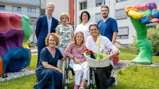Eine Gruppe von Menschen, darunter eine Frau im Rollstuhl, lächelt und posiert für ein Foto vor einem Gebäude. Ein Mann hält einen Korb voller Gegenstände.