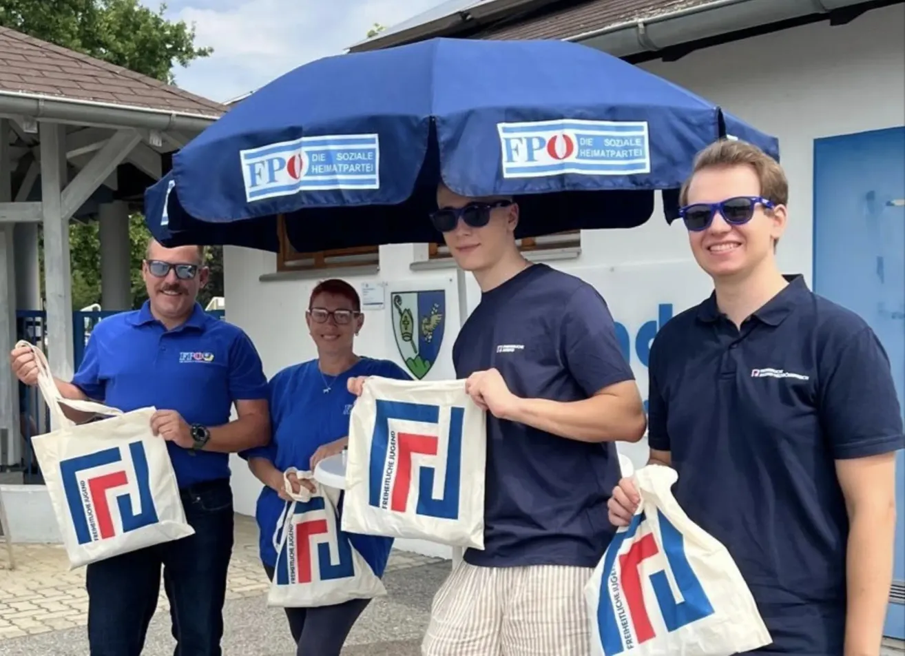 Four individuals, all dressed in blue shirts, are standing under a blue umbrella with the letters FPO on it. They are holding bags with the same letters.