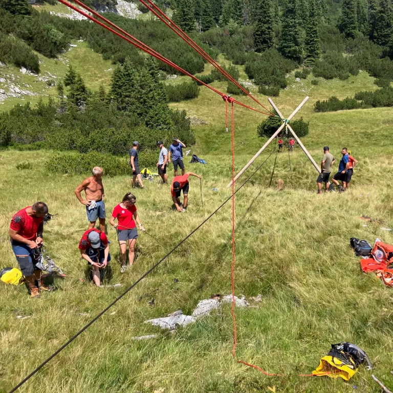 Eine Gruppe von Menschen in roten Shirts arbeitet an einer Seileinrichtung in einem Grasfeld mit einem Berghintergrund.