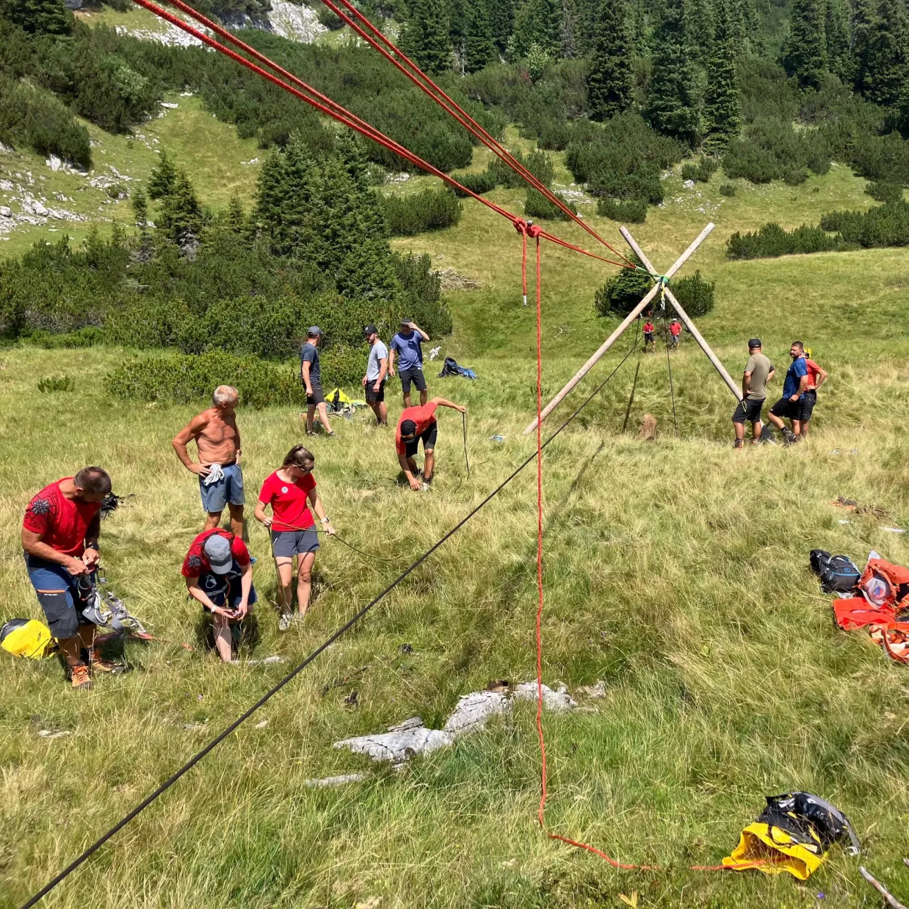 Eine Gruppe von Menschen in roten Shirts arbeitet an einer Seileinrichtung in einem Grasfeld mit einem Berghintergrund.