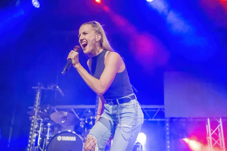 Eine Frau mit blonden Haaren singt auf der Bühne mit einem Mikrofon, trägt ein blaues ärmelloses Top und zerrissene Jeans. Hinter ihr steht ein Schlagzeug mit Yamaha-Logo.