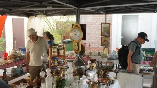 A flea market with various items including clocks, vases, and glassware under a tent. A man in a white cap is looking at the items.