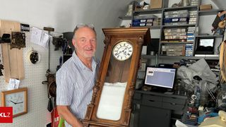 A man in a workshop holds a large wooden grandfather clock with a white face and Roman numerals. He is smiling and appears to be posing for a photo.