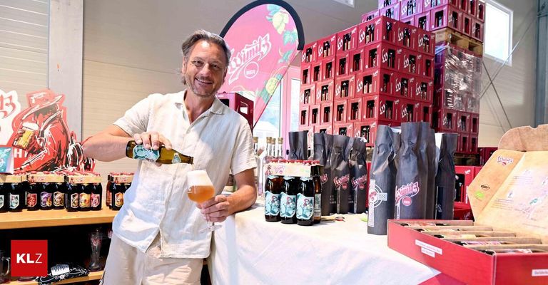 A man pours a bottle of beer into a glass at a beer tasting event, with various beer bottles and packaging in the background.