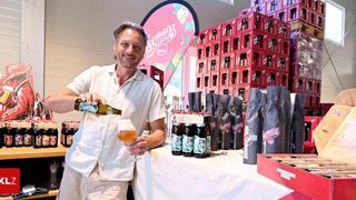 A man pours a bottle of beer into a glass at a beer tasting event, with various beer bottles and packaging in the background.