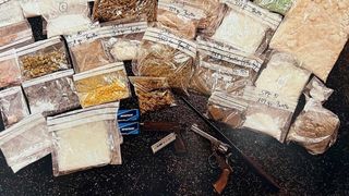 A collection of various items including plastic bags of different substances, a gun, and a box of cigarettes are laid out on a black surface. The bags are labeled with numbers and weights.
