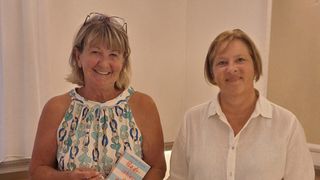 Two women standing close to each other, smiling, one holding a book titled 'Bade-schones'. The woman on the left has glasses on her head and is wearing a sleeveless blouse. The woman on the right is wearing a white shirt.
